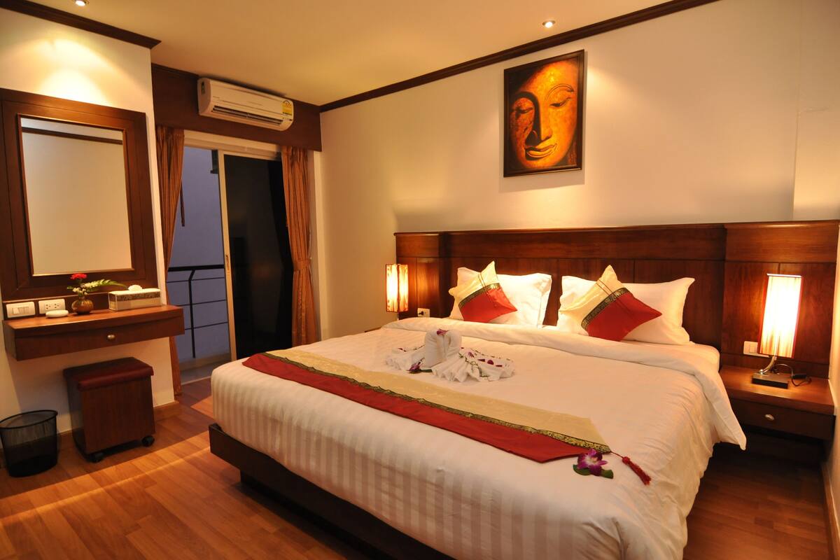 A hotel room with a large bed, a wooden headboard, a wall-mounted Buddha statue, a desk with a mirror, and a small trash bin.