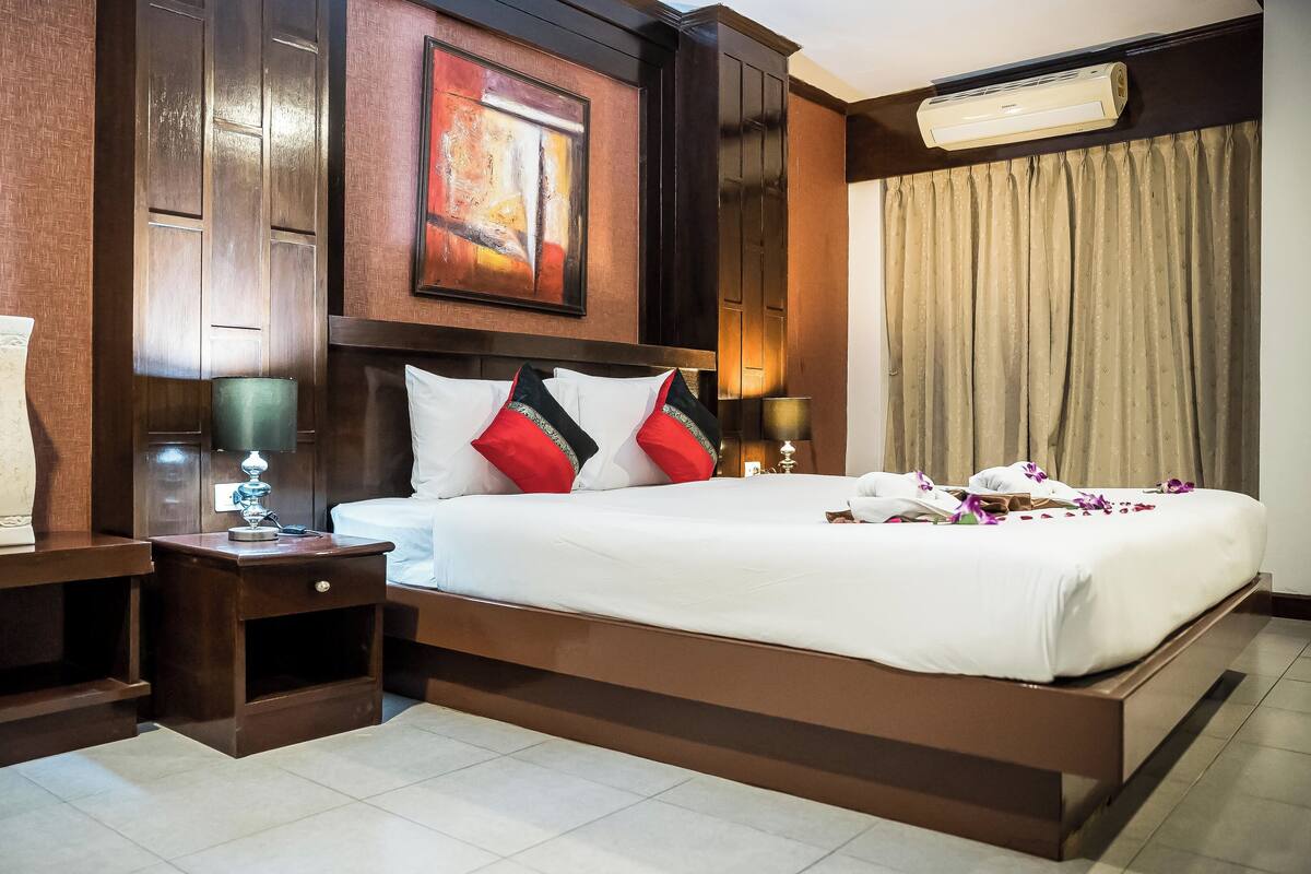 A hotel room with a large bed, a nightstand, a lamp, a painting, and an air conditioning unit.
