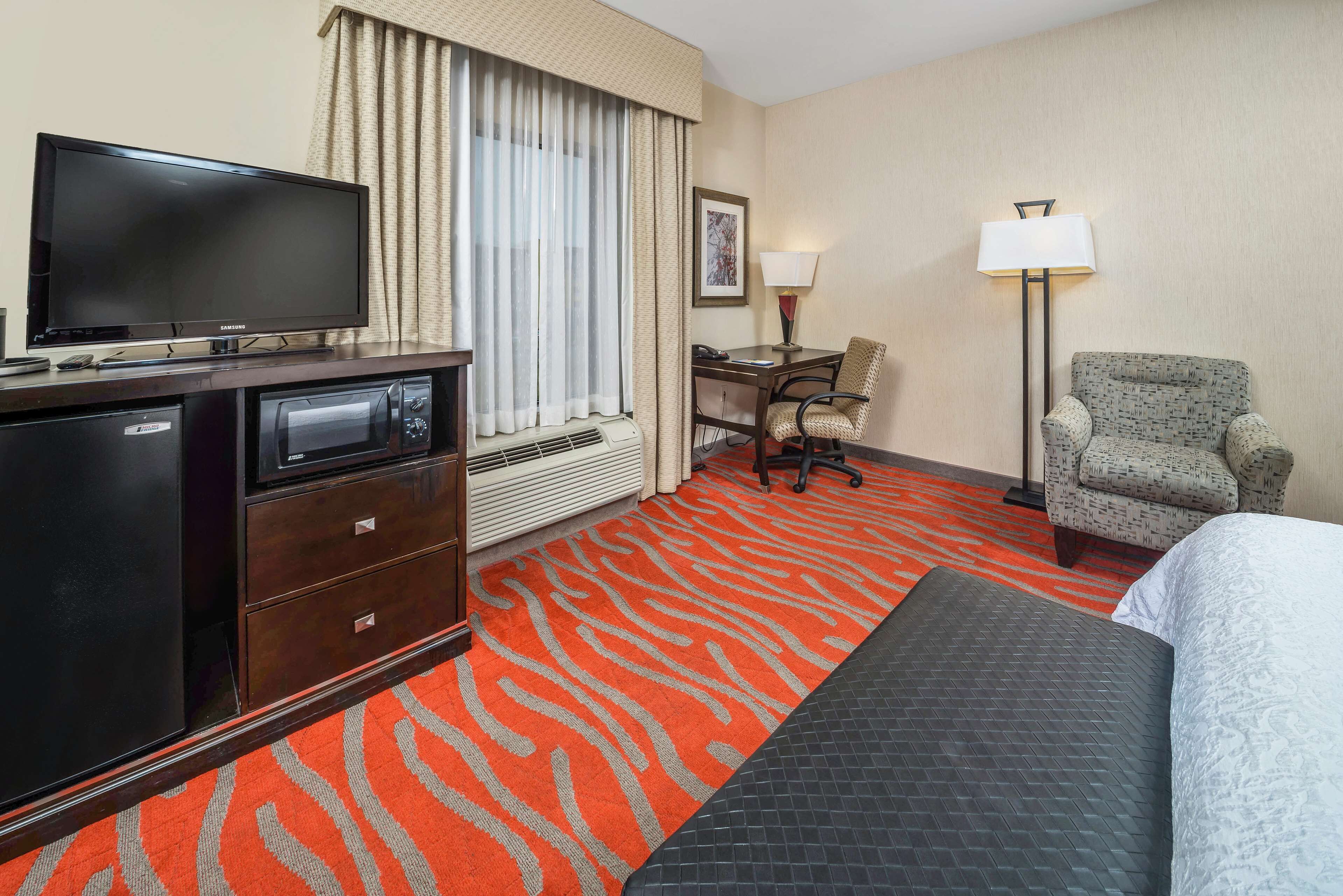 A hotel room with a flat-screen TV on a wooden cabinet, a desk with a chair, a standing lamp, and a bed with a patterned bedspread.