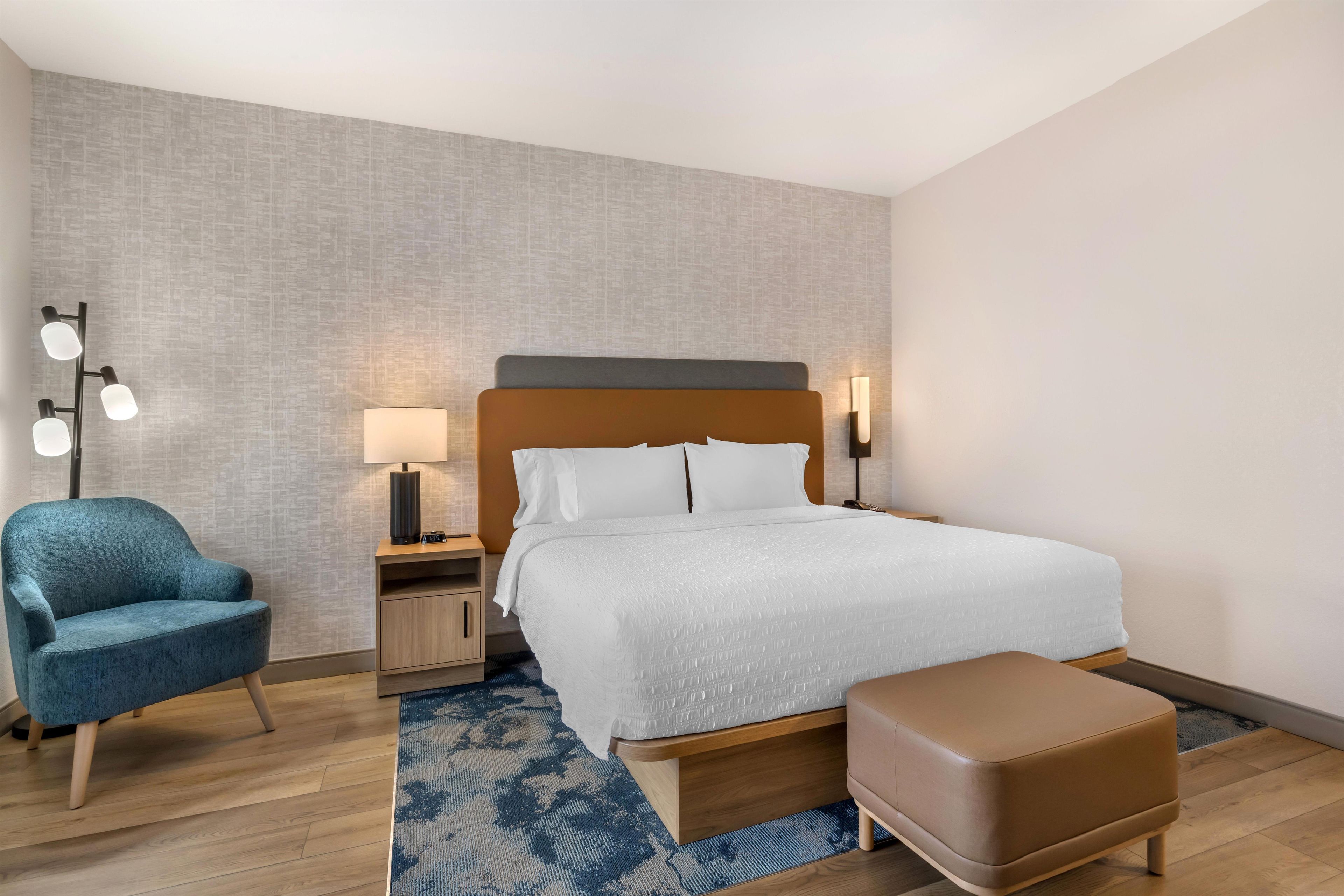 Una habitación de hotel con una cama, una silla, una mesita de noche, una lámpara y una alfombra.