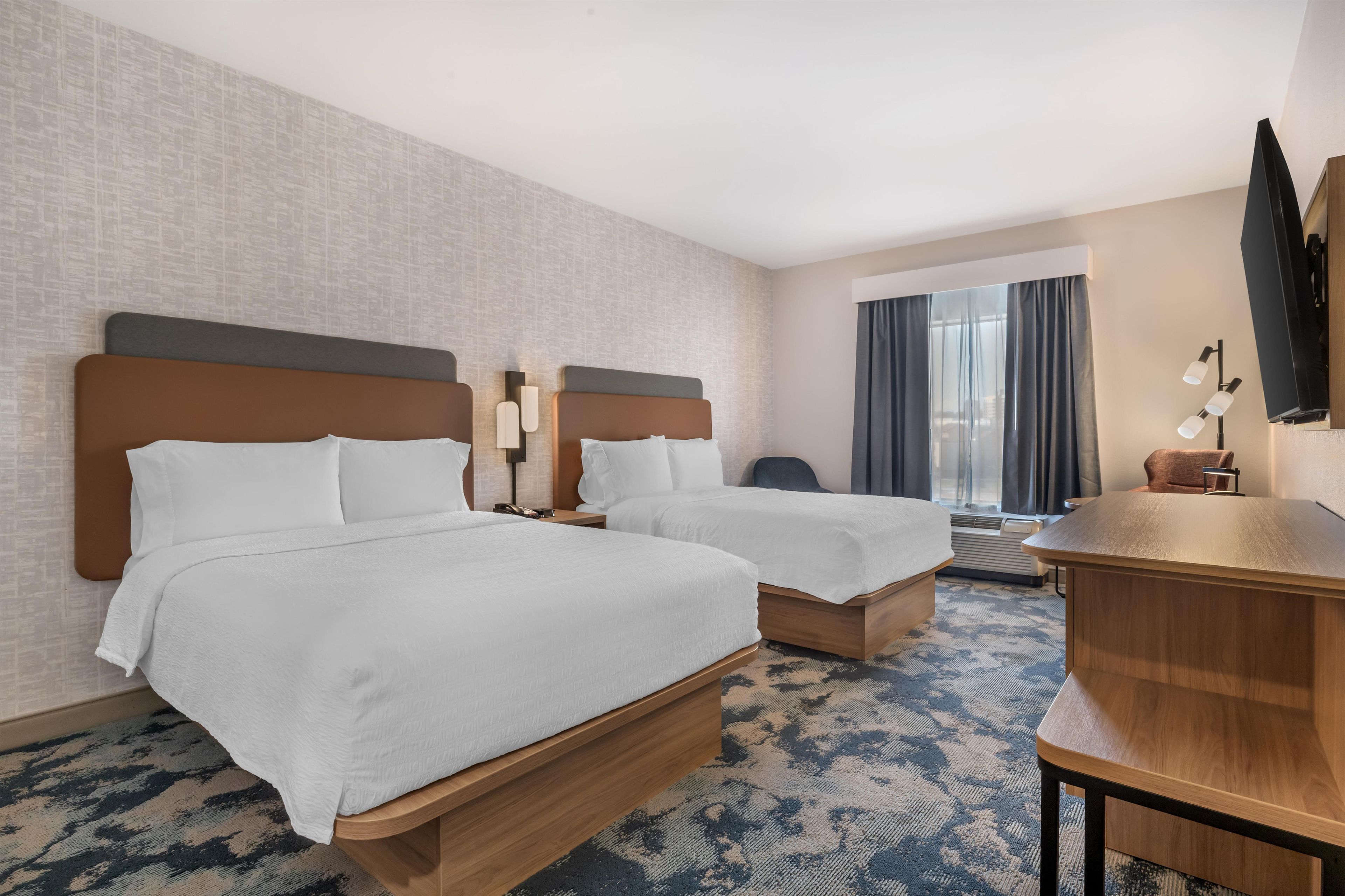 A hotel room with two beds, a desk, a chair, a TV, and a window with curtains.