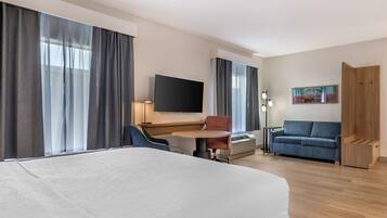 A hotel room with a bed, a desk with a chair, a sofa, a TV mounted on the wall, and a painting.