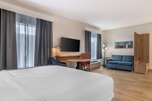 A hotel room with a bed, a desk with a chair, a sofa, a TV mounted on the wall, and a painting. - Hampton Inn & Suites Tulsa/Catoosa (Catoosa)