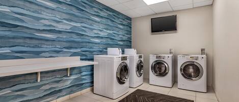 A laundry room with a tiled floor, a bench, three washing machines, a flat-screen TV on the wall, and a patterned rug.