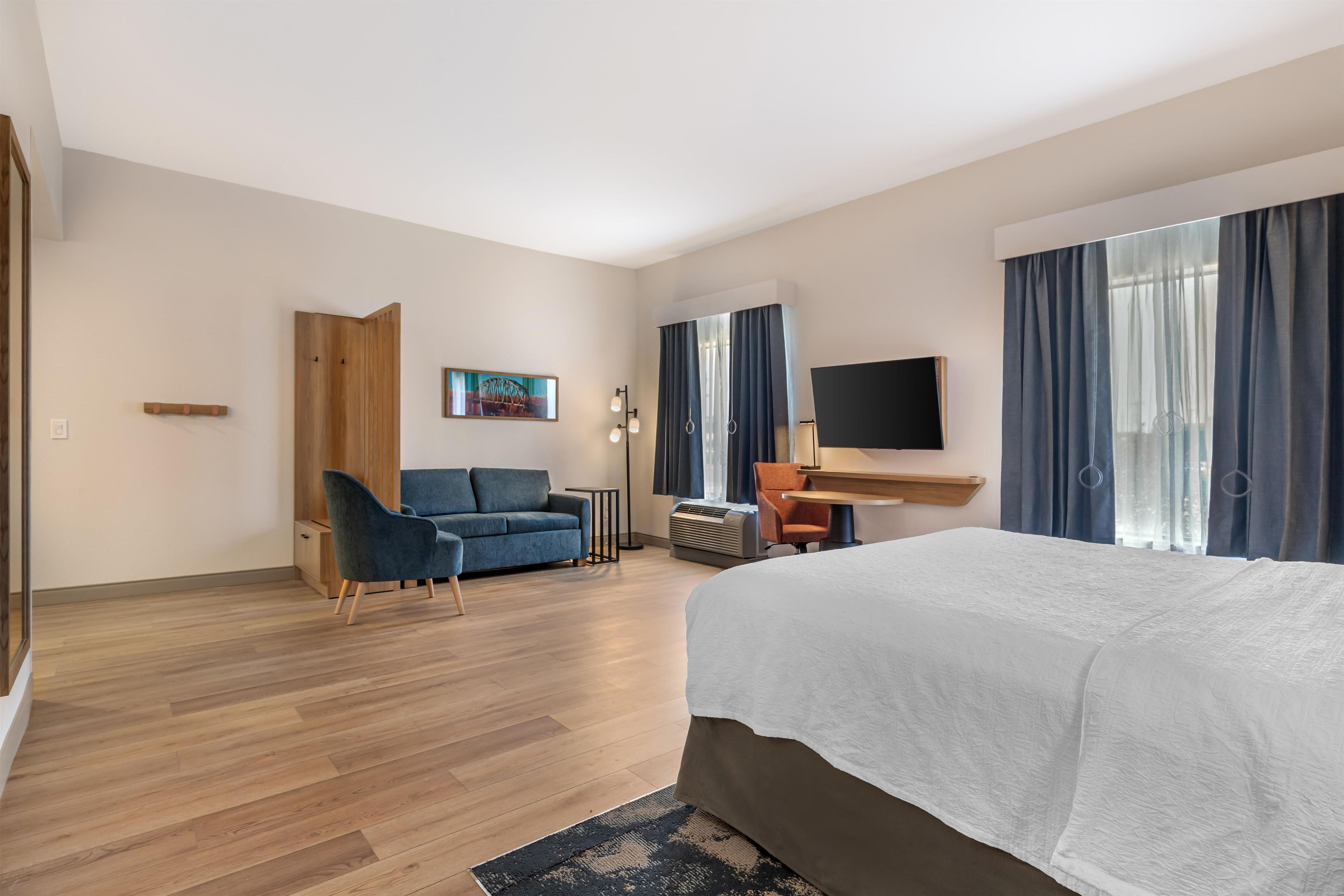 A hotel room with a bed, a sofa, a desk, a TV, and a painting on the wall.