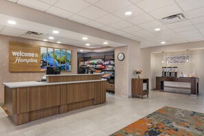 A hotel reception area with a wooden reception desk, a clock, and a wall-mounted welcome sign. - Hampton Inn & Suites Tulsa/Catoosa (Catoosa)
