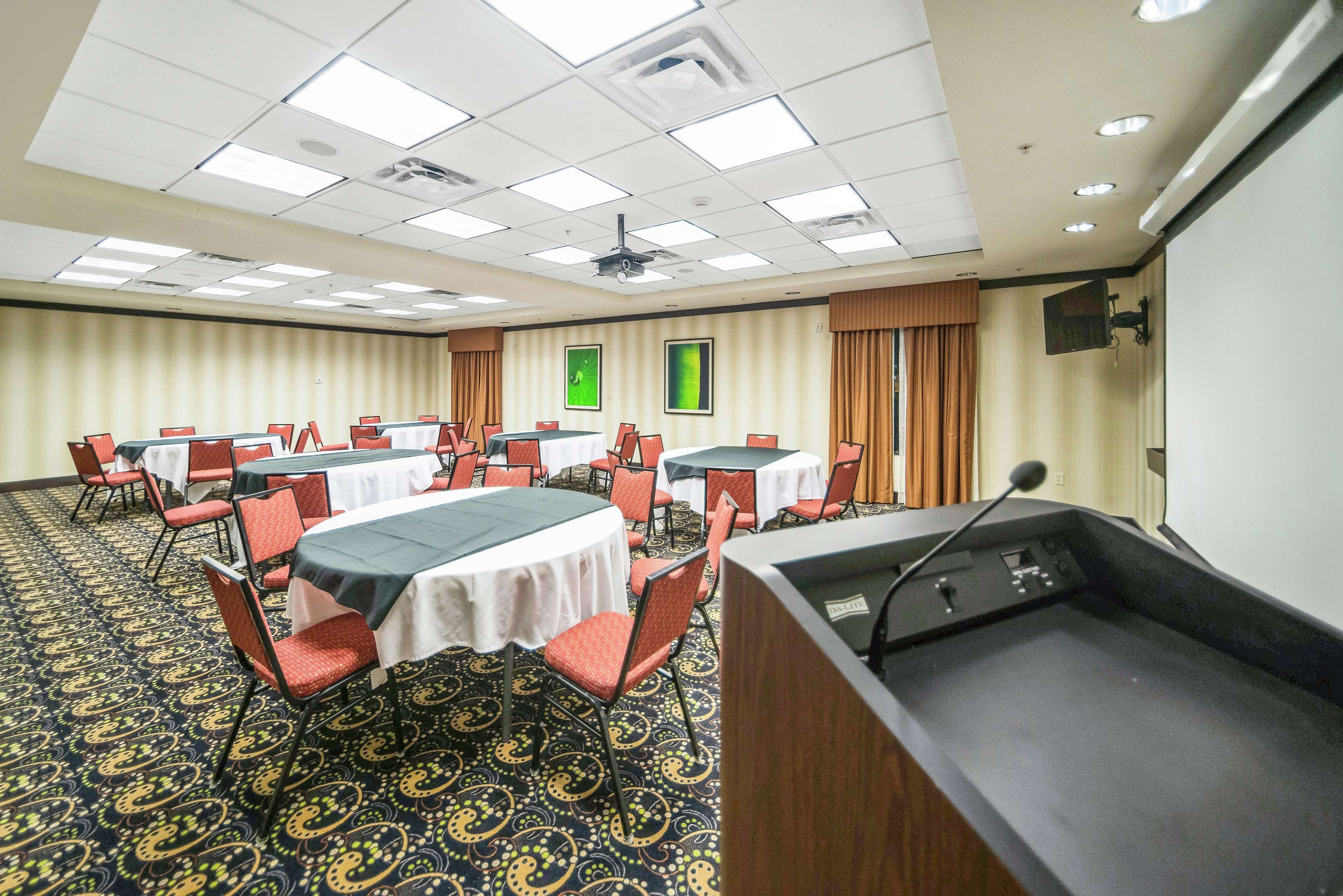 Un salón de conferencias con varias mesas y sillas, un atril y una pantalla de proyector.