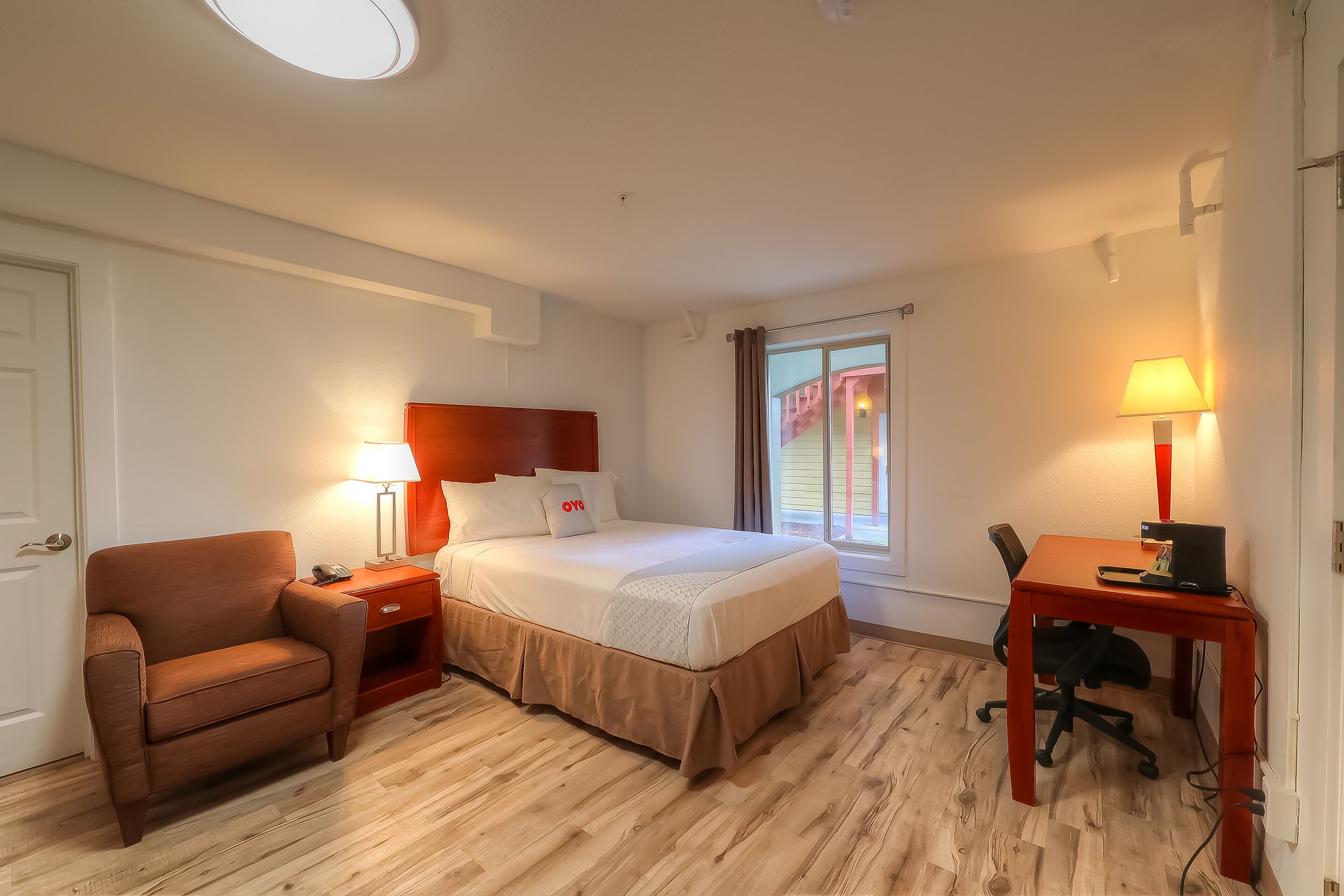 A hotel room with a bed, a desk, a chair, a nightstand, and a lamp.