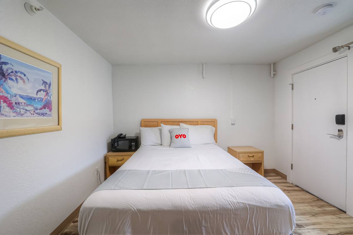 Une chambre d’hôtel bien rangée, avec un lit, des tables de chevet, une petite table, un tableau encadré au mur et une porte.