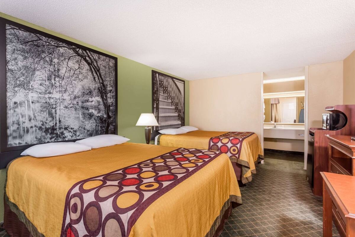 A hotel room with two beds, a desk, a chair, a lamp, a microwave, and a large framed picture on the wall.