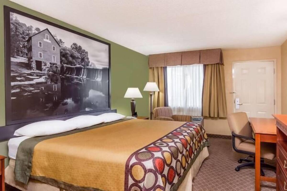 A hotel room with a bed, a desk, a television, a chair, a lamp, a window with curtains, and a painting on the wall.