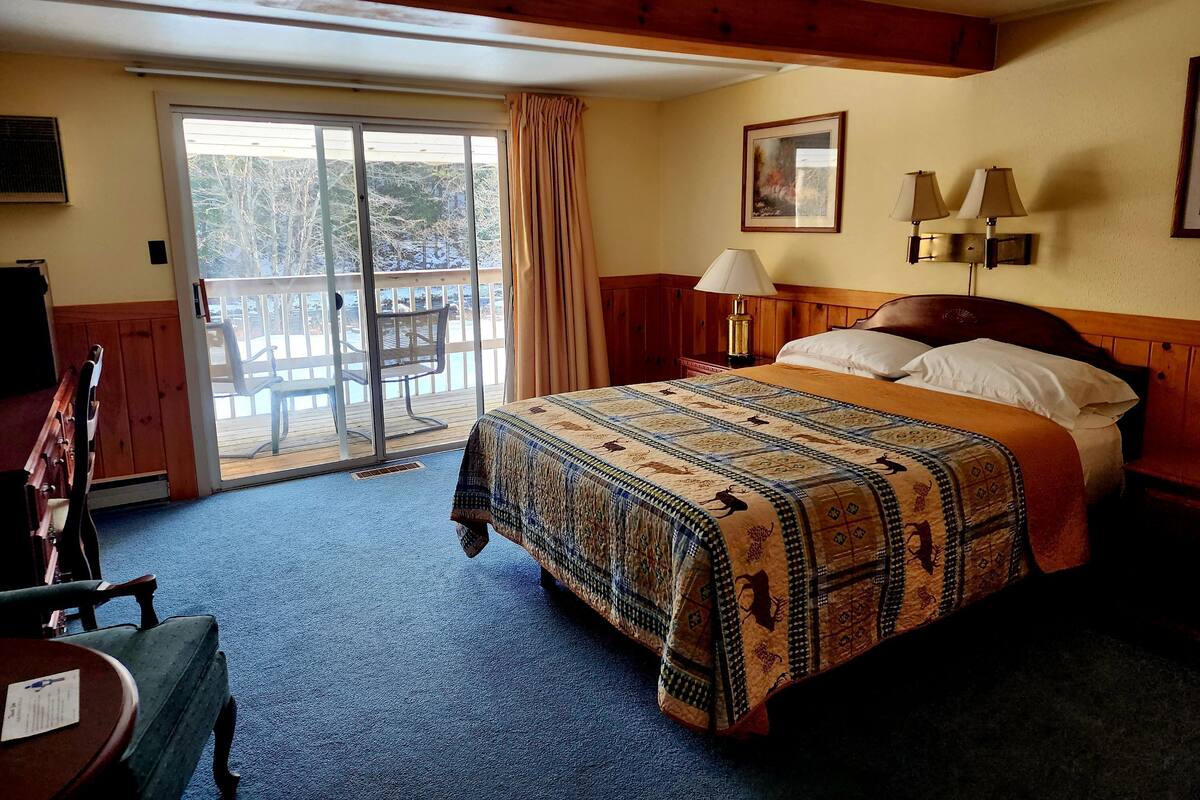 A hotel room with a large bed, a desk with a chair, a balcony with a chair, and a painting on the wall.