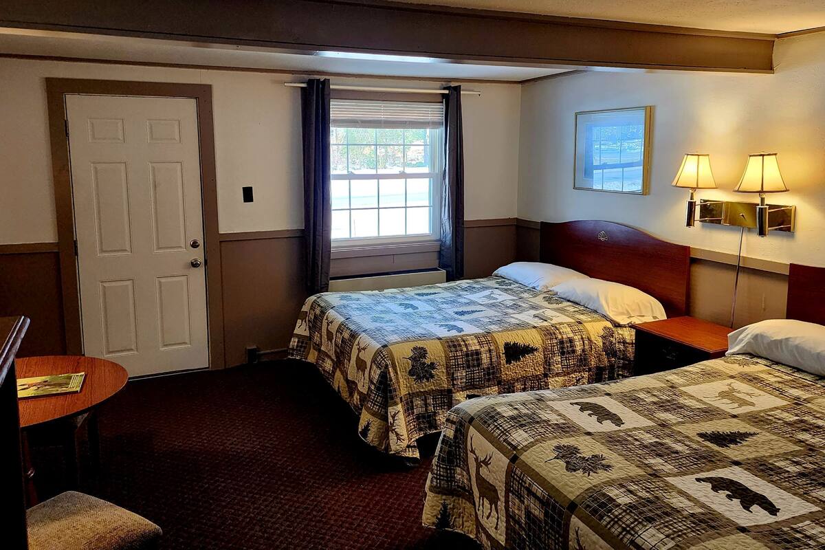 A hotel room with two beds, a door, a window with curtains, a wall-mounted lamp, and a framed picture.