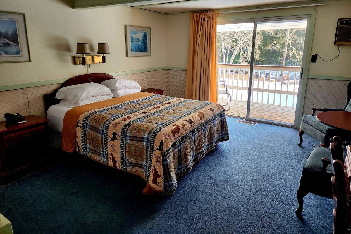 A hotel room with a bed, a desk, a chair, a balcony with a view, and two wall-mounted lamps.