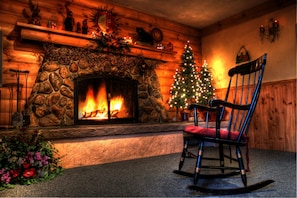 A cozy fireplace with a lit fire, a decorated Christmas tree, and a rocking chair in a log cabin-style room. - Covered Bridge Riverview Lodge (Jackson)