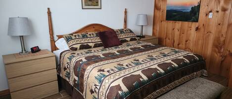 A bedroom with a wooden headboard, a bed with a patterned blanket, bedside lamps, a nightstand with a clock, a wooden bench, and a wall-mounted picture.
