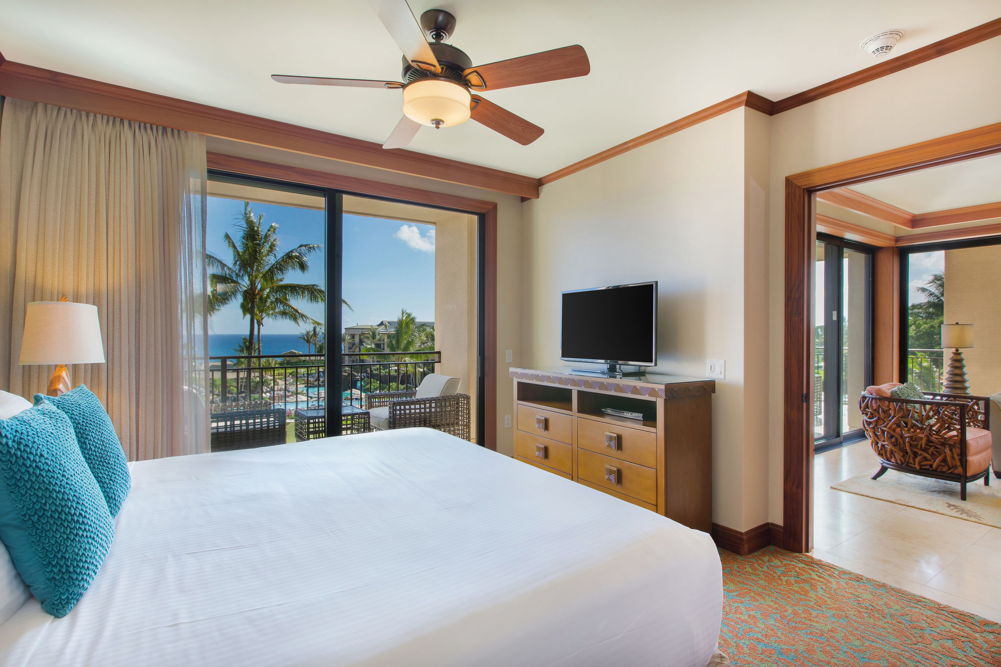 A hotel room with a large bed, a television, a balcony with a view of palm trees, and a ceiling fan.