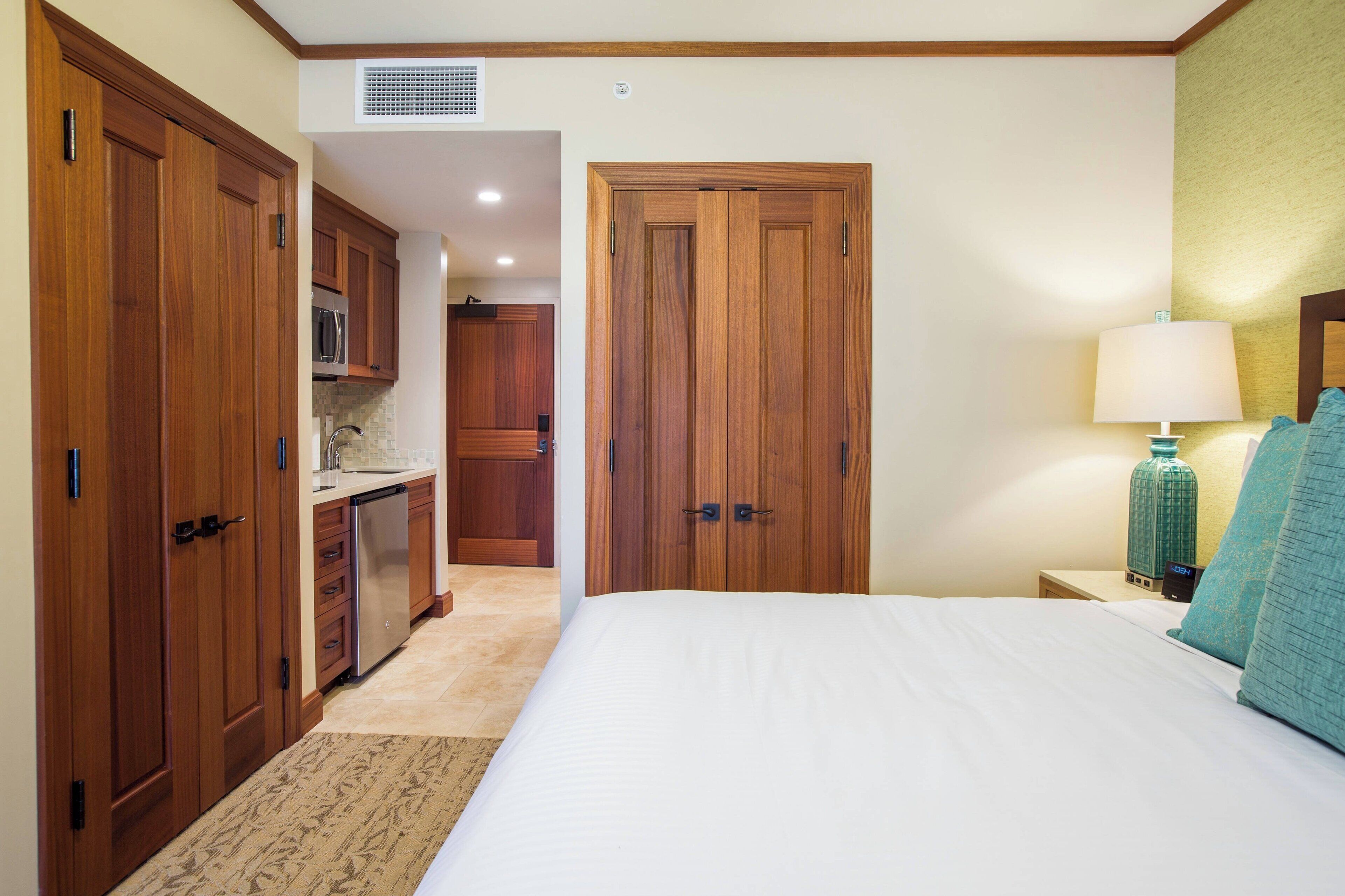 A bedroom with a bed, a nightstand, a lamp, and a view of a kitchen area.