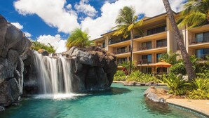 A hotel with a pool featuring a waterfall, palm trees, and a clear blue sky. - Koloa Landing Resort at Poipu, Autograph Collection (Koloa)