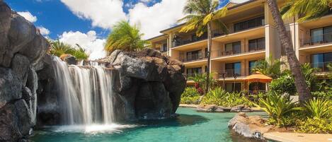 Un hotel con piscina que incluye una cascada, palmeras y un cielo azul despejado.