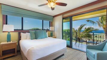 A hotel room with a large bed, a ceiling fan, a balcony with a view of palm trees and the ocean, and a seating area with a chair.