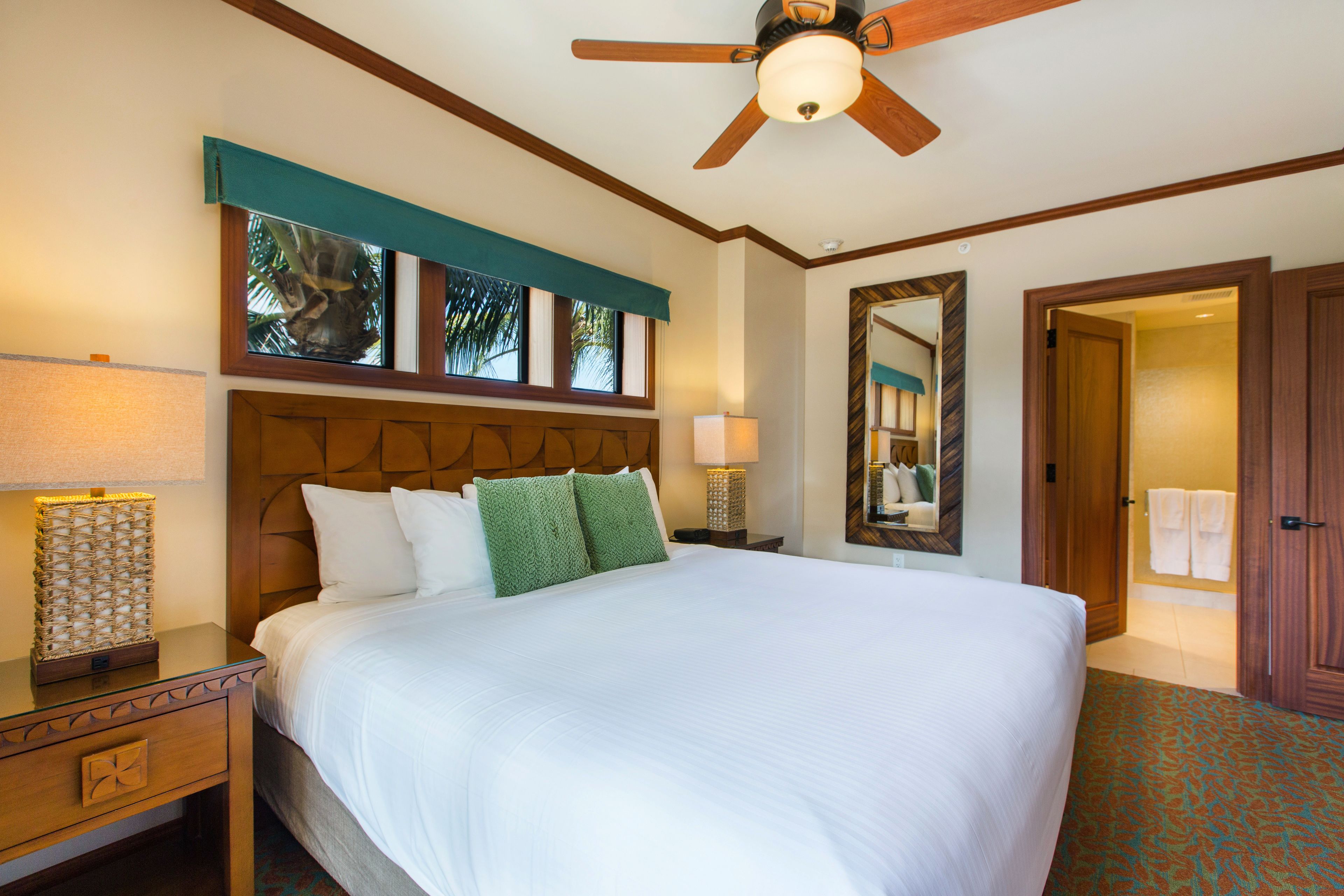 Une chambre d’hôtel avec un grand lit, une tête de lit en bois, deux lampes de chevet, un ventilateur de plafond et un grand miroir au mur.