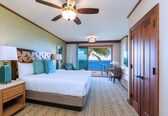 A hotel room with a bed, a nightstand, a ceiling fan, a view of the ocean, and a balcony with a chair.