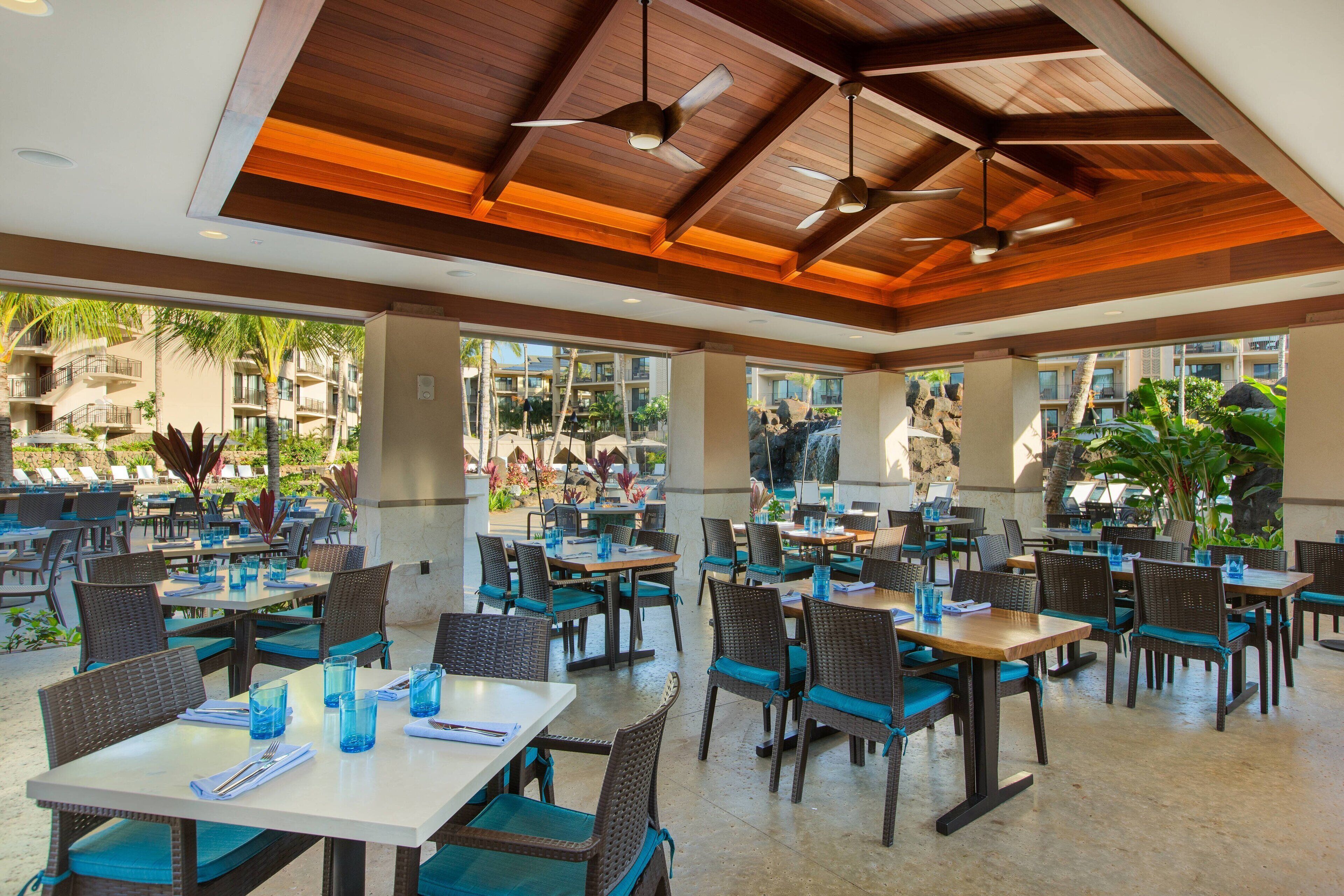 An outdoor dining area with tables and chairs, large windows, and a view of the surrounding buildings and greenery.