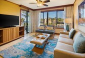 A hotel room with a large flat-screen TV, a sofa, a coffee table, and a balcony with a view of palm trees and buildings.