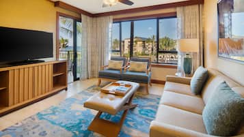 Habitación de hotel con televisor de pantalla plana grande, sofá, mesa de centro y balcón con vistas a palmeras y edificios.