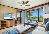 A spacious living room with a television, comfortable seating, a ceiling fan, and large windows offering a view of the outdoors.