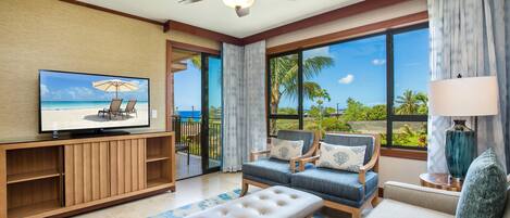 A spacious living room with a television, comfortable seating, a ceiling fan, and large windows offering a view of the outdoors.