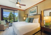 A hotel room with a large bed, a ceiling fan, a balcony with a view of palm trees, and a painting on the wall.