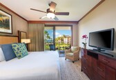 A hotel room with a bed, a television, a ceiling fan, a chair, a dresser, and a balcony with a view of palm trees and buildings.