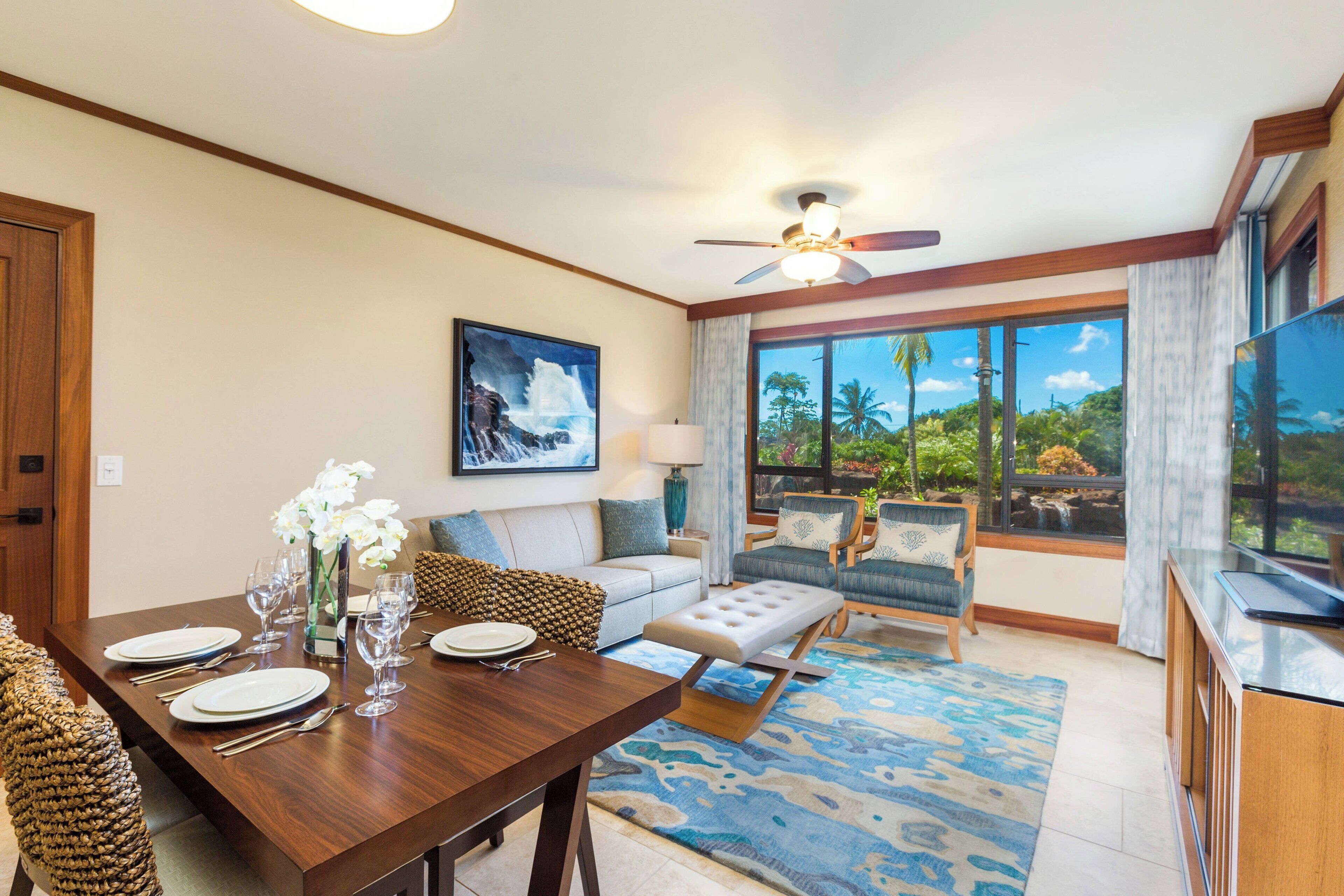 A spacious living room with a dining area, a sofa set, a coffee table, and a large window offering a view of palm trees and a waterfall.