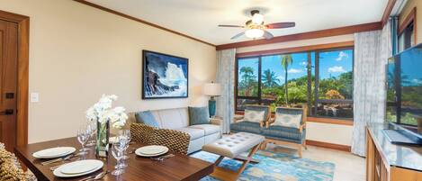 A spacious living room with a dining area, a sofa set, a coffee table, and a large window offering a view of palm trees and a waterfall.