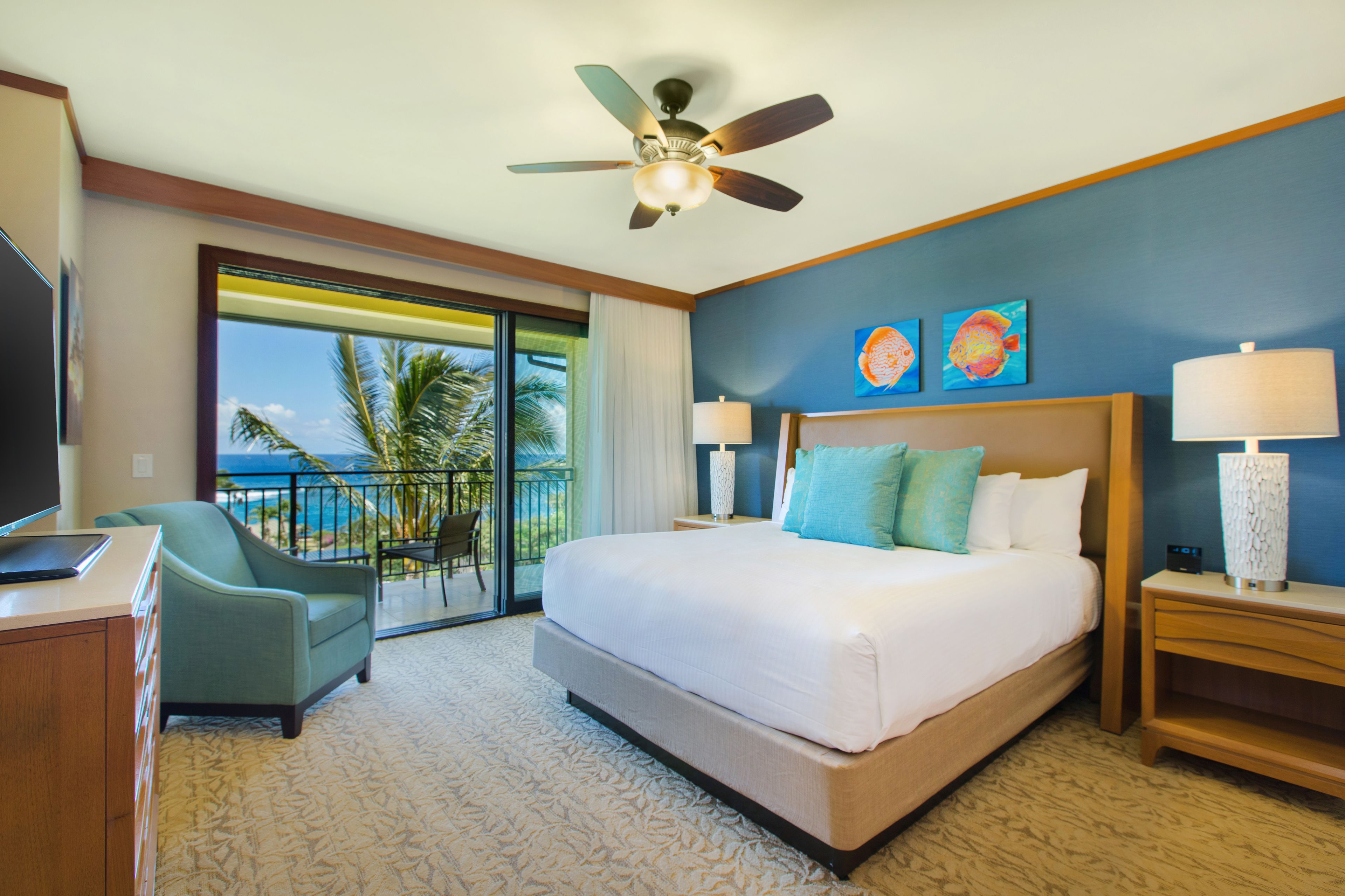 A hotel room with a large bed, a ceiling fan, a desk with a computer, a chair, and a view of the ocean through a sliding door.