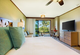 A hotel room with a large bed, a television, a balcony with a view of palm trees, and a dining area with a table and chairs.