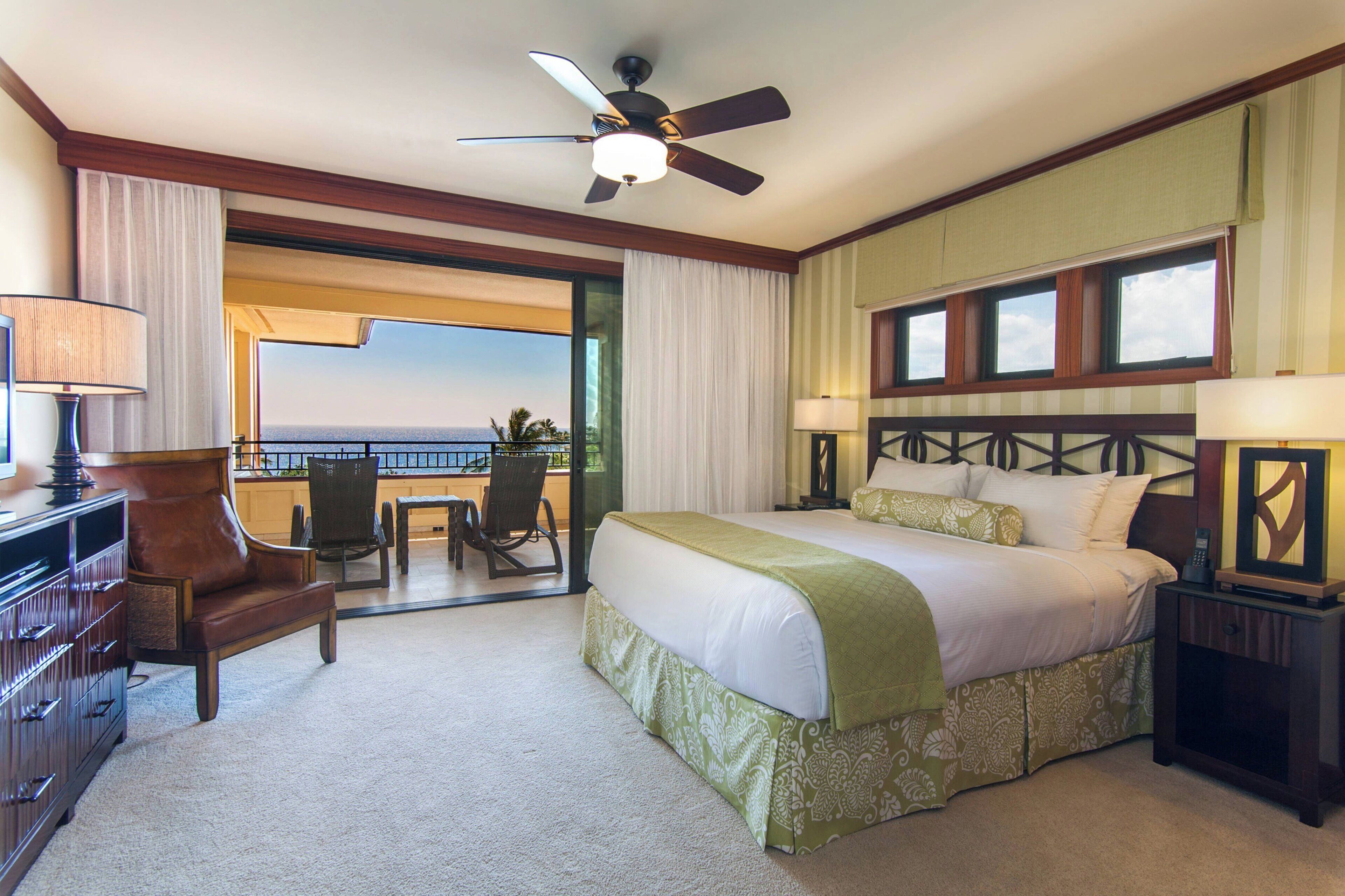 Habitación de hotel con cama, escritorio, silla, ventilador de techo y vista al océano.