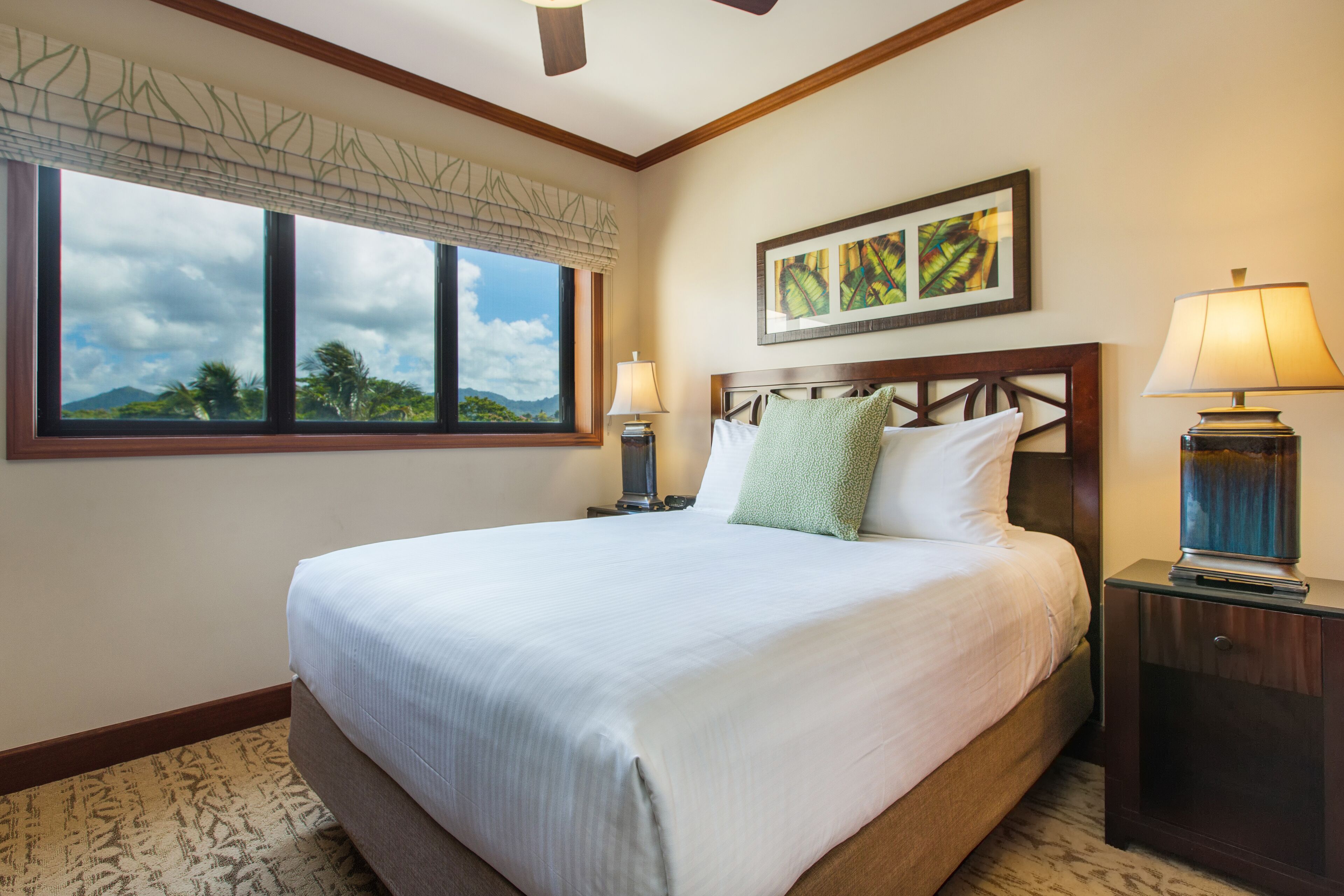 A neatly made bed with a headboard, a nightstand with a lamp, a window with a view of the outdoors, and a framed picture on the wall.