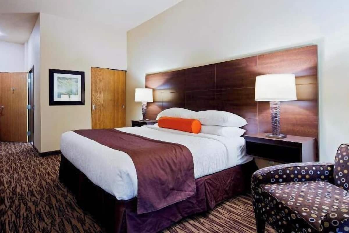A hotel room with a bed, two bedside tables with lamps, a TV, a desk, and a patterned chair.