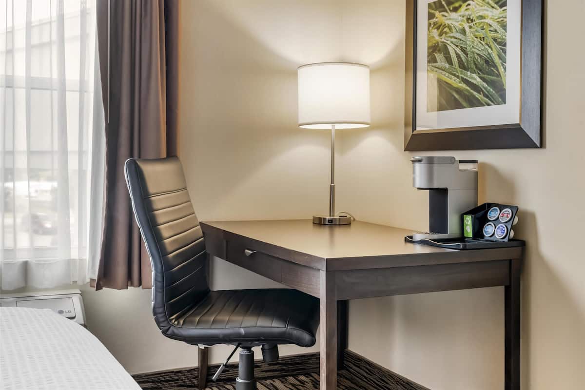 A hotel room with a desk, chair, lamp, coffee machine, and a framed picture on the wall.