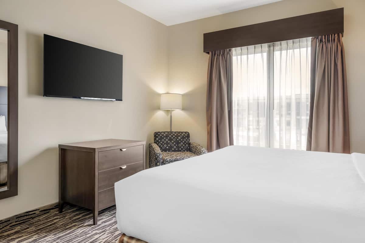 A hotel room with a bed, a TV mounted on the wall, a desk with drawers, a chair, and a window with curtains.