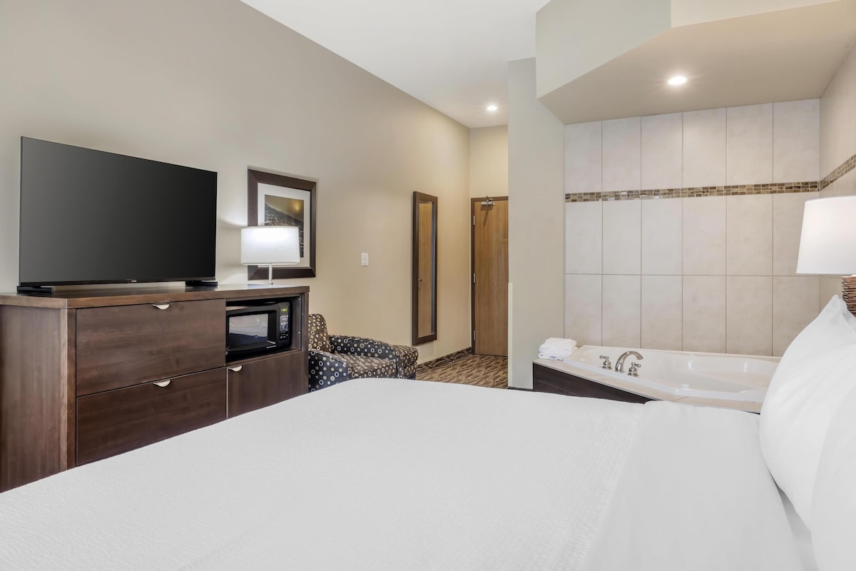 Habitación de hotel con una cama grande, televisor de pantalla plana, escritorio con microondas, zona de estar con sillones estampados, baño con bañera y pared con azulejos.