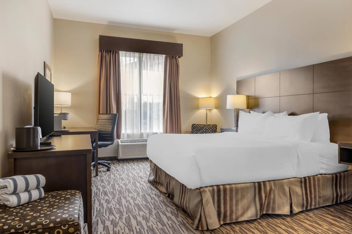 A hotel room with a large bed, a desk with a TV, a chair, a small table with a lamp, and a window with curtains.