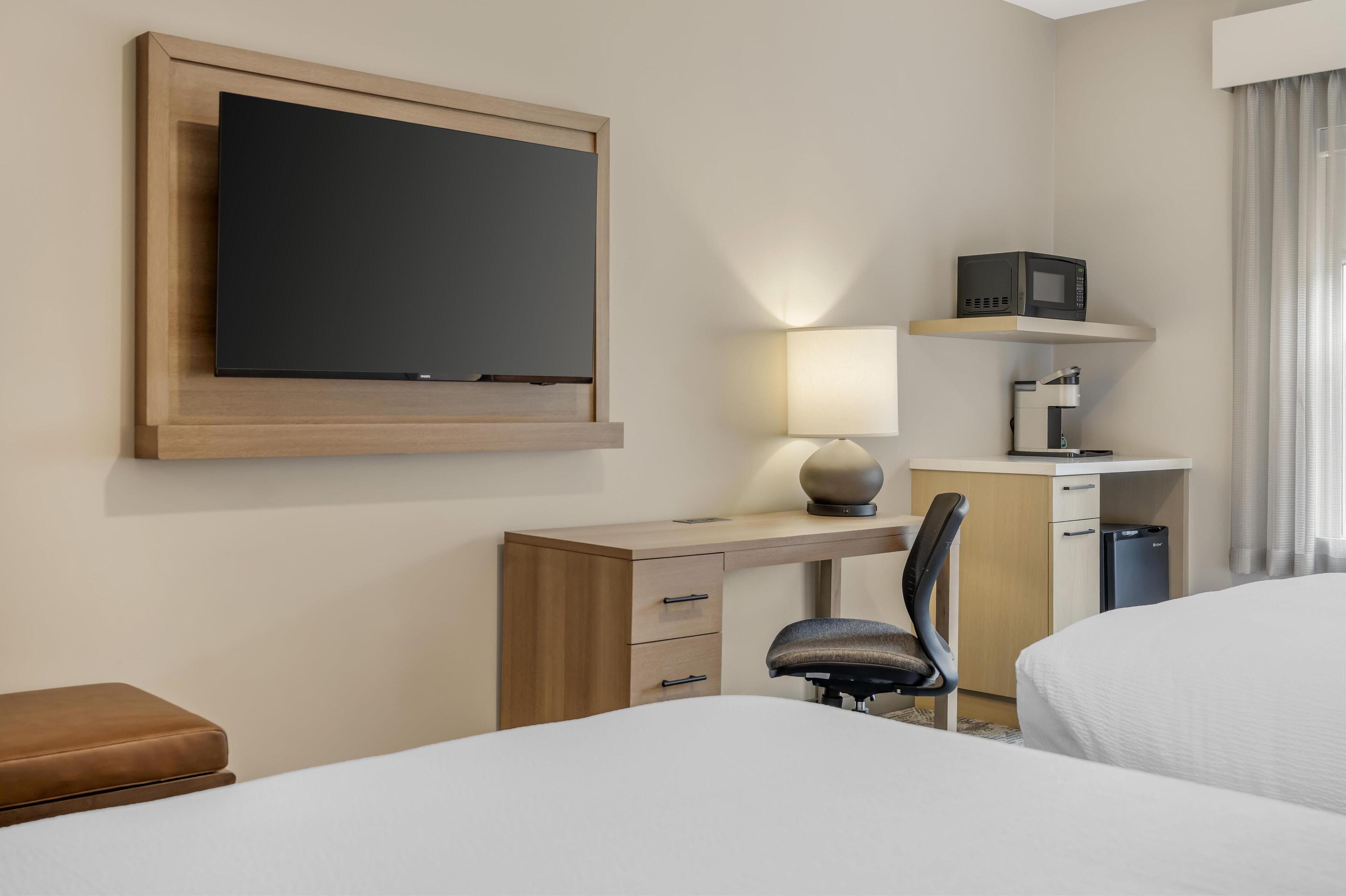 Een hotelkamer met een bed, een bureau met stoel, een aan de muur bevestigde televisie, een lamp, een magnetron en een koelkast.