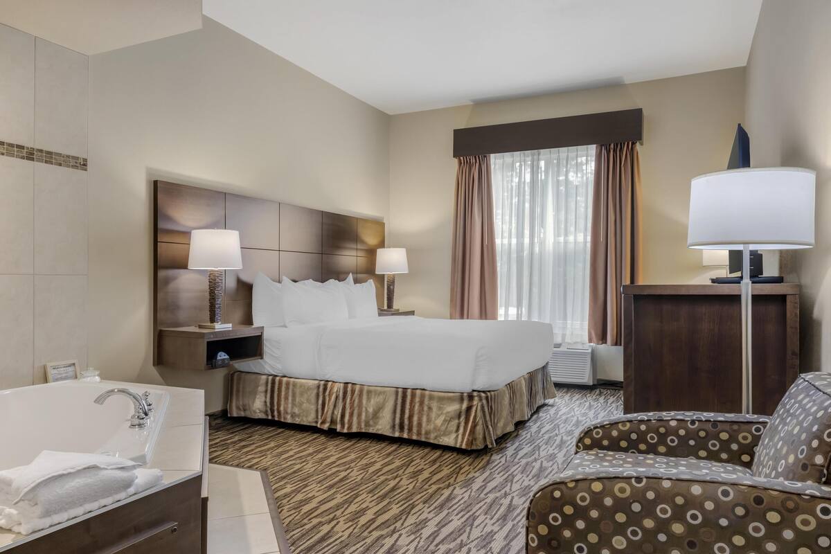 A hotel room with a large bed, two bedside lamps, a wooden headboard, a large window with curtains, a patterned carpet, a sofa, and a bathroom with a bathtub and towel.