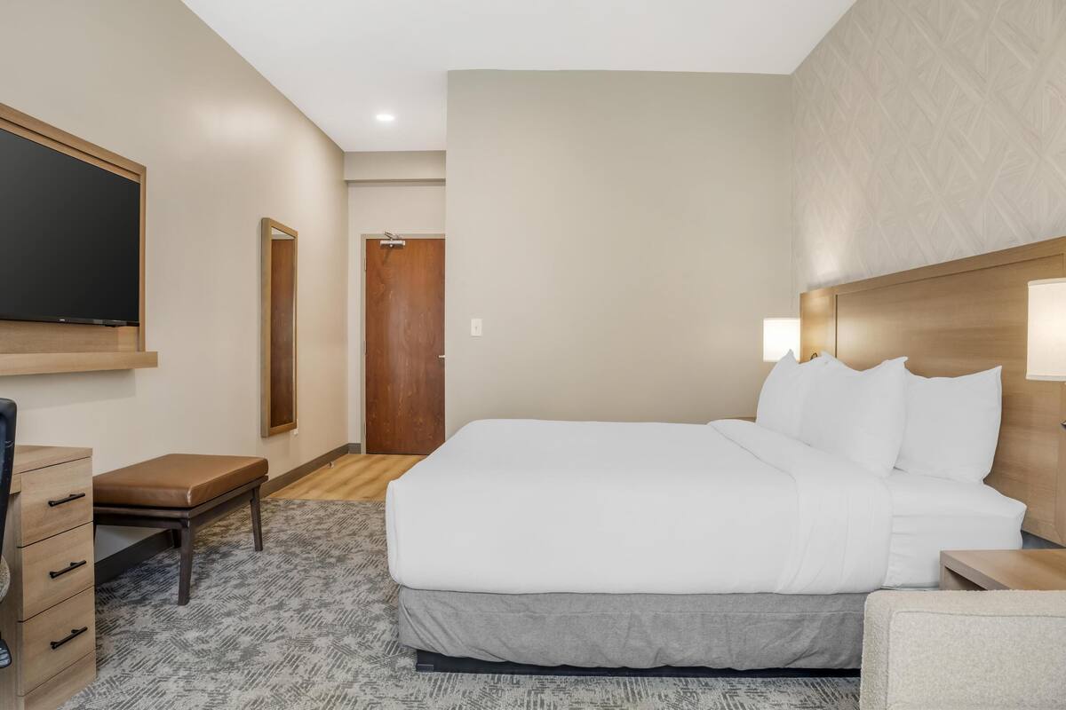 A hotel room with a large bed, a desk with drawers, a chair, a TV mounted on the wall, and a door leading to another room.