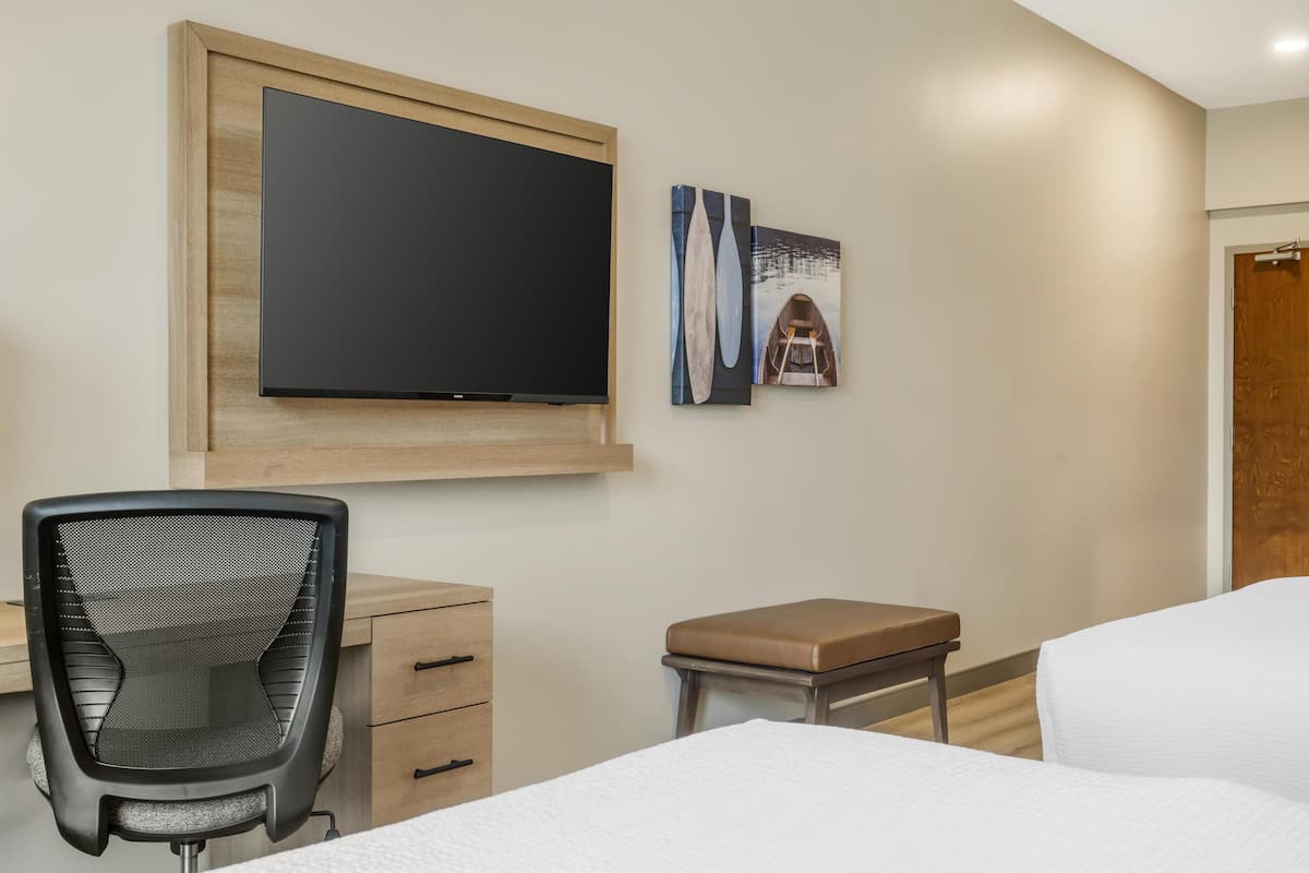 A hotel room with a bed, a desk with a chair, a television mounted on the wall, and a small table with a bench.