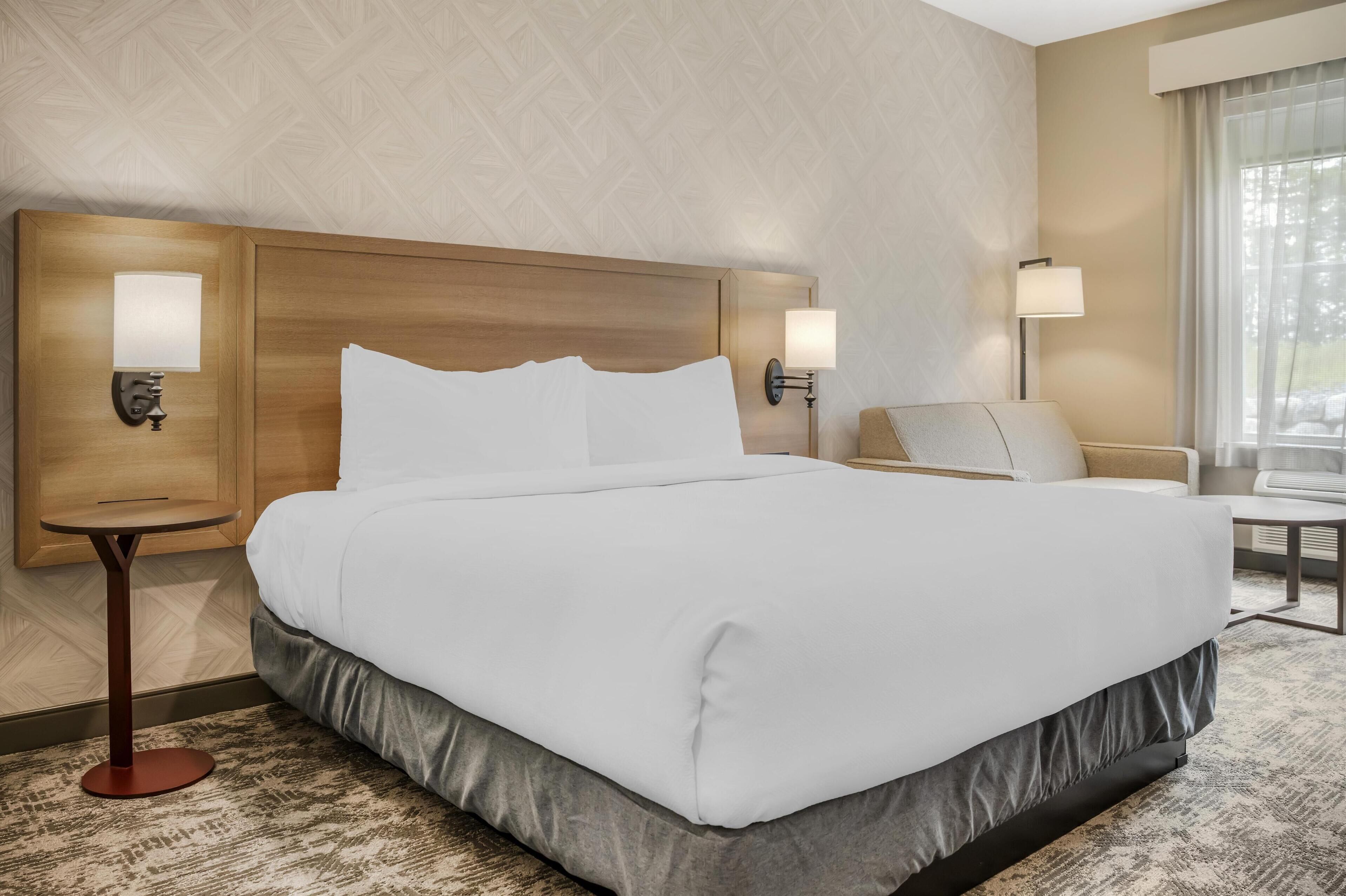 A hotel room with a large bed, two bedside lamps, a small table, and a chair.